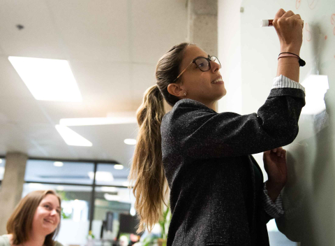 An instructor writes on a whiteboard while a student looks on.