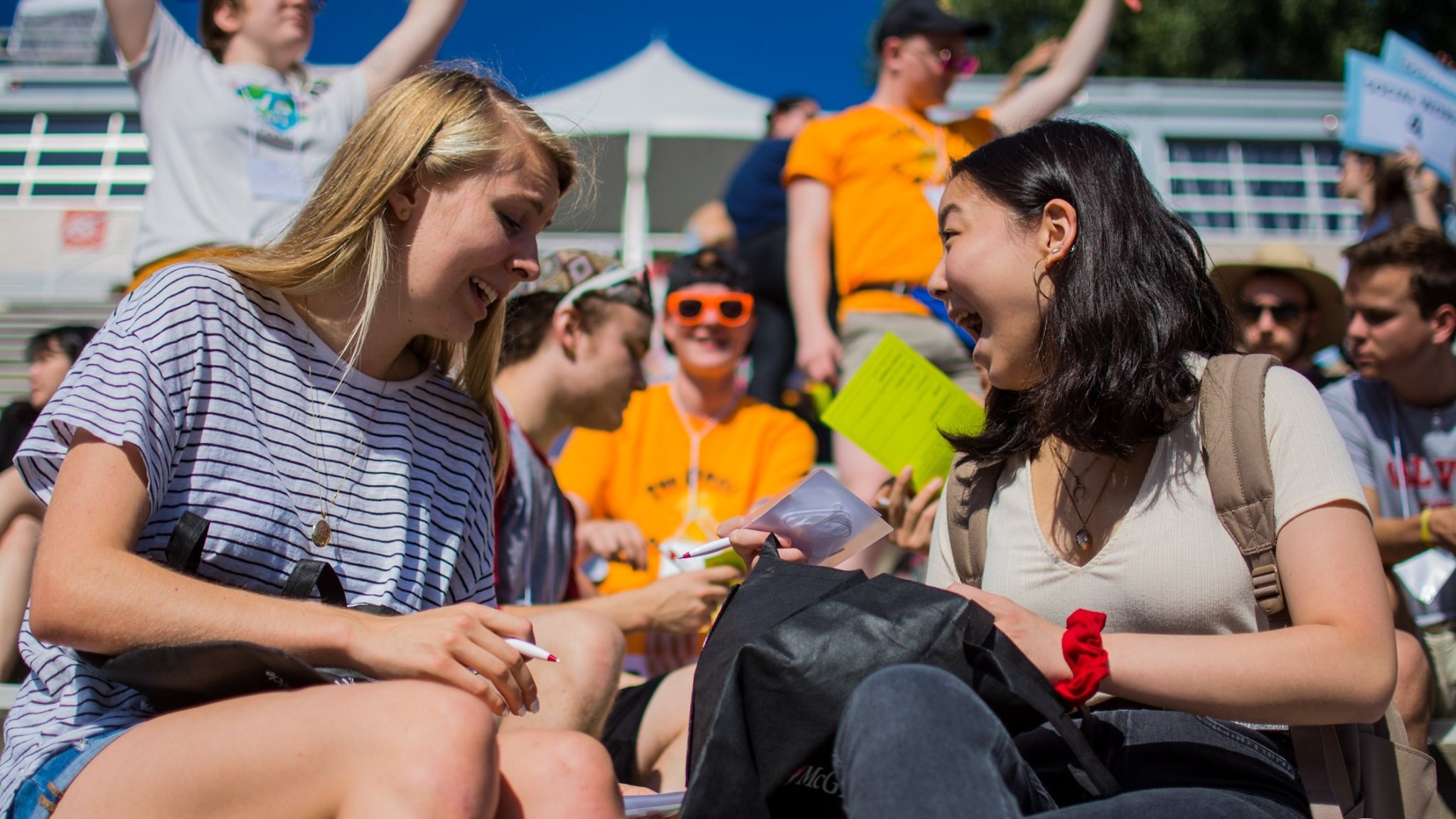 a blonde student and a dark-haired student smile joyfully while sitting in a crowd in the sun