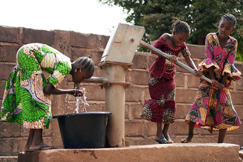 One young girl drinking from a public well while two other girls pump water