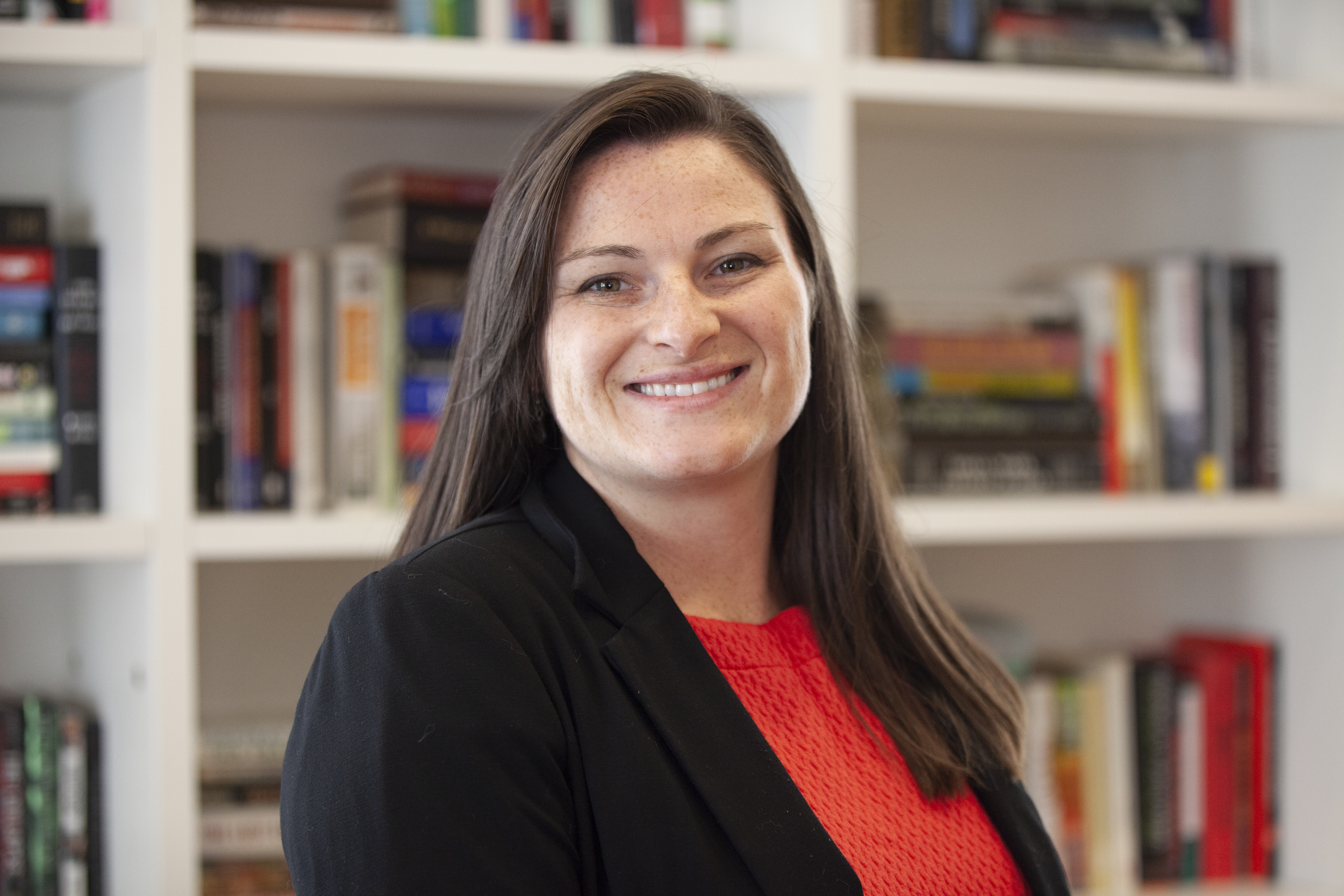 Marin MacLeod - woman with straight dark shoulder-length hair wearing a black jacket and red blouse posing in front of bookshelves