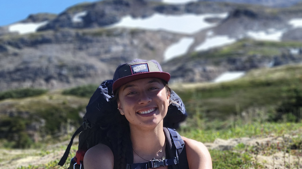 Allyson Kis - Young woman posing in front of a mountain top wearing a tank top, hiking back pack and baseball cap