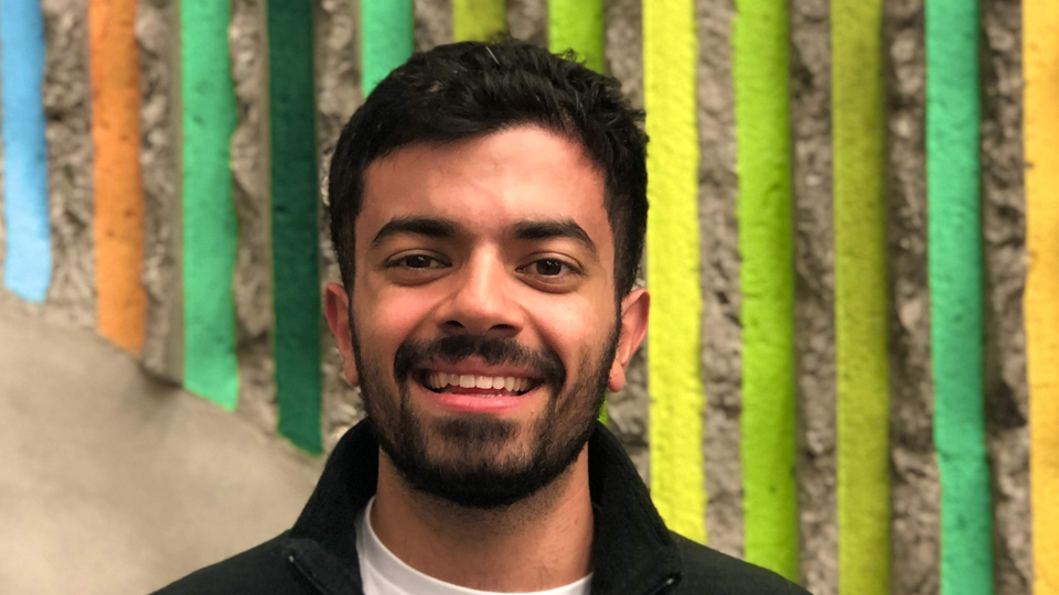 headshot of Cedric Kafie - young man with dark hair and beard posing in front of a colourful wall