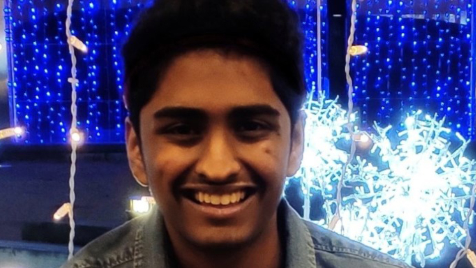 Maahir Shah - smiling young South Asian man wearing a denim jacket posing in front of a blue and white light installation