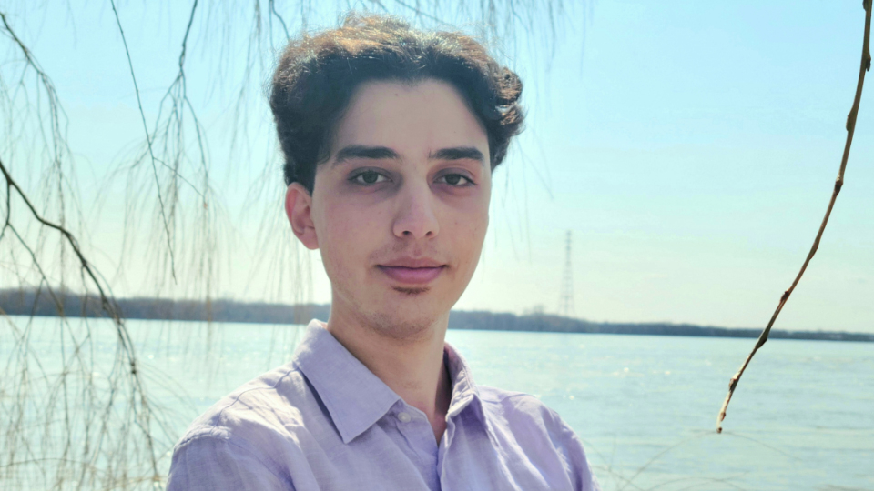 Mehrshad Bakhshi - young man with short wavy dark hair and light purple shirt standing in front of a lake