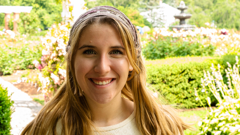 headshot of Sadie Valente - young woman with light brown hair and a headband posing in front of a garden