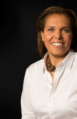 Dr. Gina Ogilvie - woman with shoulder length hair wearing a white button-down shirt and a chunky necklace posing in front of a dark background