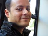 un homme aux cheveux courts vous regarde. Sourire, vêtements sombres. Il est dans le bus