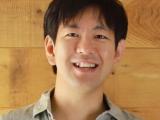 A young Japanese postdoc is standing against a wooden wall. Broad smile, cheerful attitude.