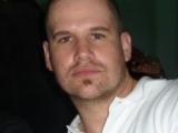 un homme portant une chemise blanche est représenté sur cette photo. Pas de cheveux, regard direct.