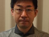 A Japanese neurosurgeon wearing glasses and a stripy shirt looks in to the camera attentively