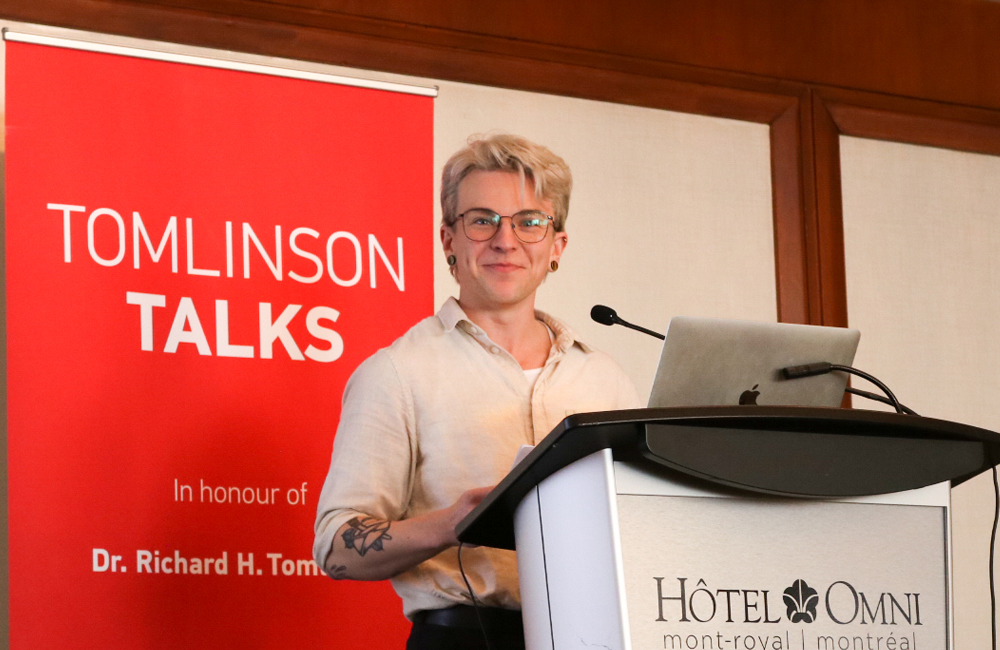 Kit is a white person with blonde hair and tattoos on their arms. They are wearing a beige shirt and glasses. They stand in front of a silver and black podium and laptop, from which they are smiling at the camera.