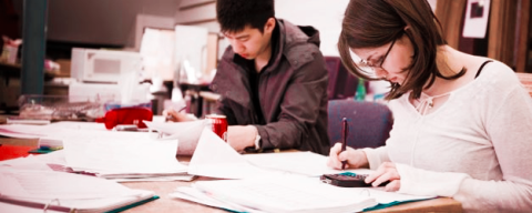 Two students review papers and books spread out on a table.