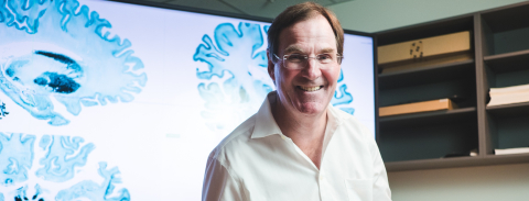 McGill Professor Alan Evans in a white shirt smiling at the camera in an office settings with an render of a brain scan in the background.