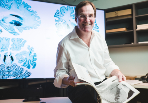 Alan Evans sits in front of blue-tinted scans of the BigBrain on a screen, holding printed black and white scans in his hands