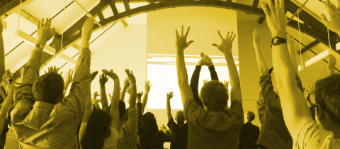 Attendees of an event stand with their arms up in the air in front of a screen with a presentation on it