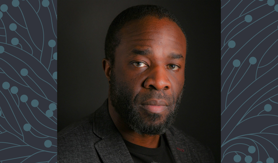 Headshot portrait of Emmanuel Olarewaju a black man with short-cropped hair, a short-trimmed beard and brown eyes, wearing a dark grey blazer over a black t-shirt, set against a dark background