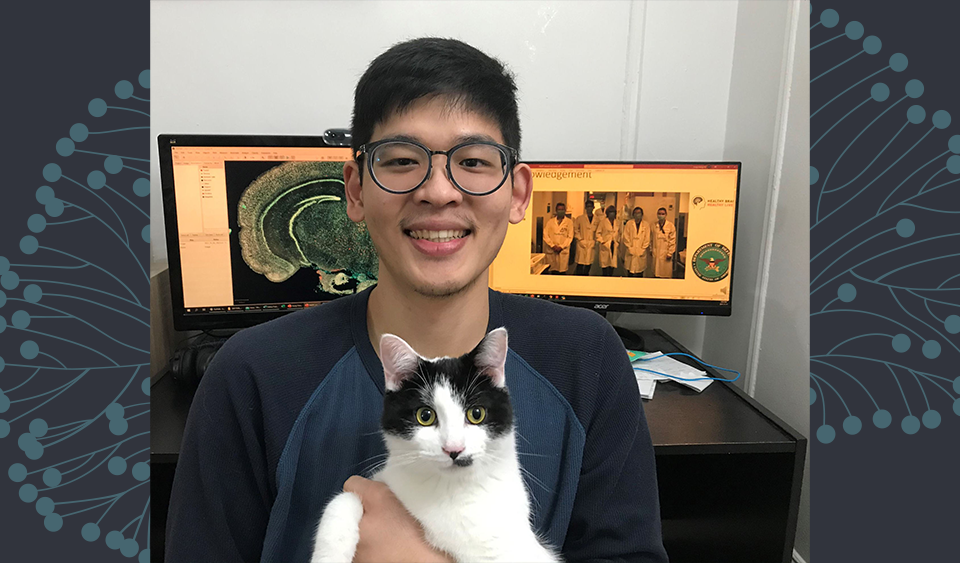 Hung-Hsiang Ho posing in front of his computer with a black and white cat.