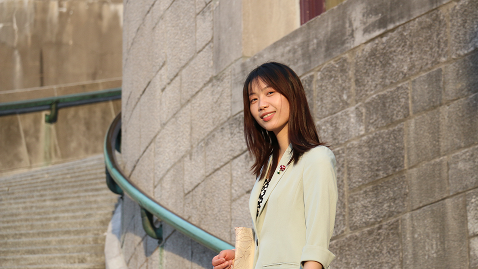 An Asian woman with brown hair, wearing a beige blazer over a black shirt with white blotches, stands on a stone stairway in front of a stone wall equipped with a green handrail