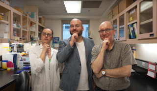 Three researchers stand in a row in a wet-lab space; they look on pensively, each with a closed fist on their chin