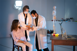 Two Piece of Mind Collective members dressed as doctors examine another member playing the role of patient