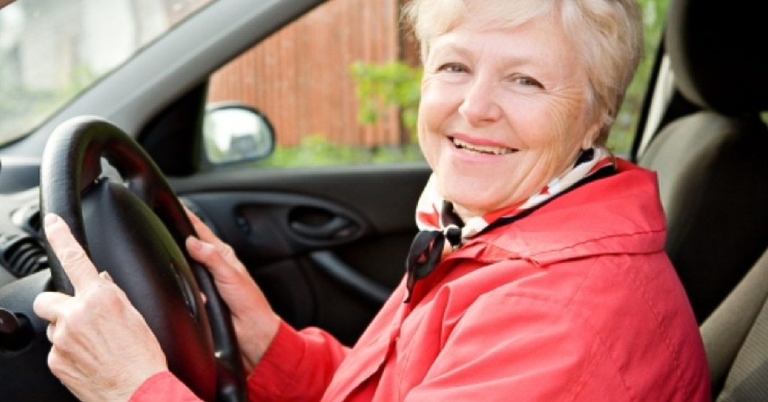 A woman in a red coat sits behind the wheel of a car and smiles. 
