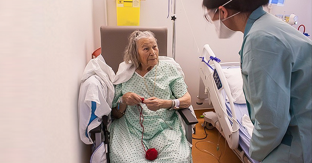 An older female patient knits next to her hospital bed while chatting to a young female physiotherapy student, who wears scrubs and a mask
