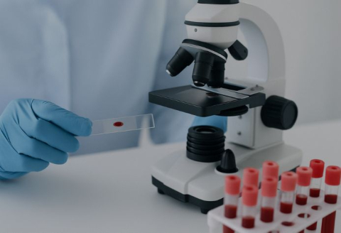 Gloved hand preparing a blood sample slide for analysis under a microscope, with blood vials in the foreground, representing education, diagnostics, and laboratory research in hematology.