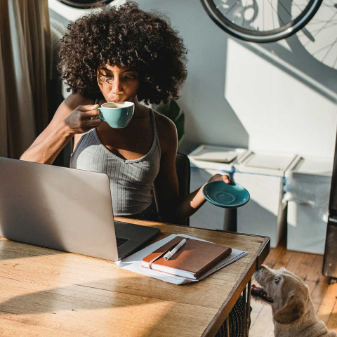 woman working on a desk and drinking coffee in front of her laptop and dog