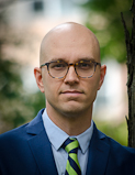 Sébastien Jodoin. Photo by Lysanne Larose. [a white bald man with glasses, wearing a pale blue shirt, a navy jacket and a tie stands next to a tree]