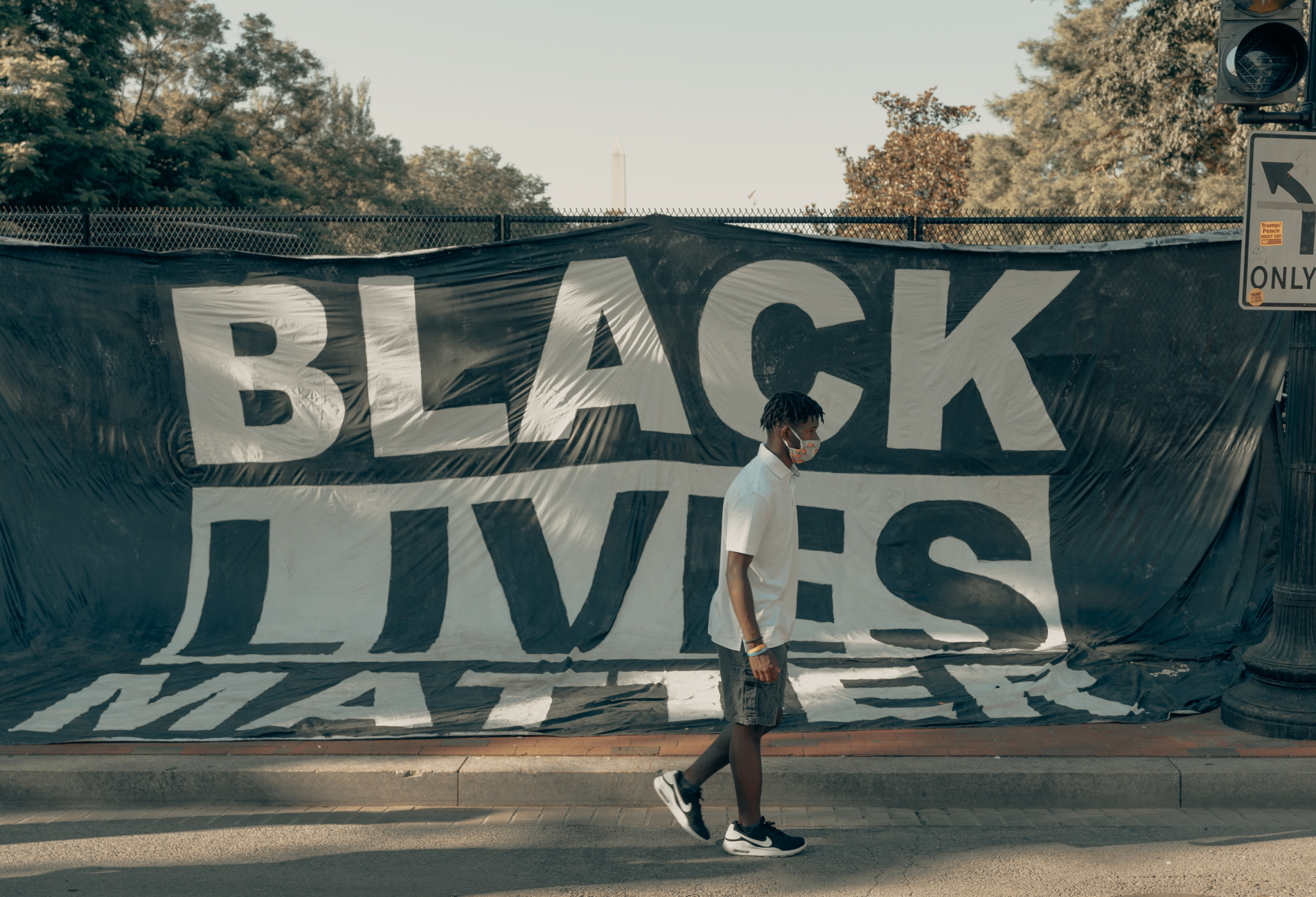 A young black men is walking on the sidewalk, behind him you see a large black and white flag reading 'Black Lives Matter'