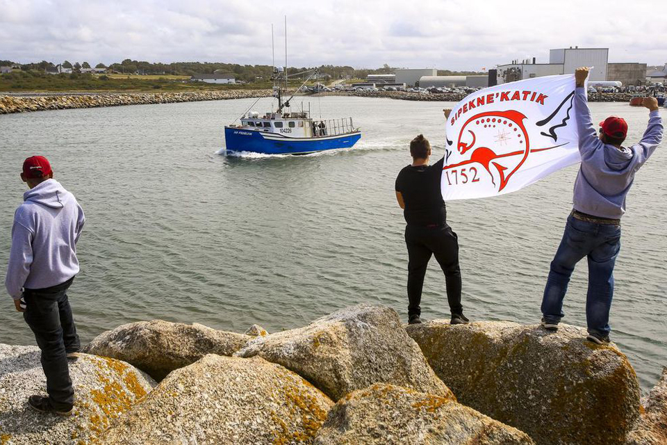 Three protesters standing on a seawall at the entrance of a harbour. A blue ship sails by. THE CANADIAN PRESS/Mark O’Neill 