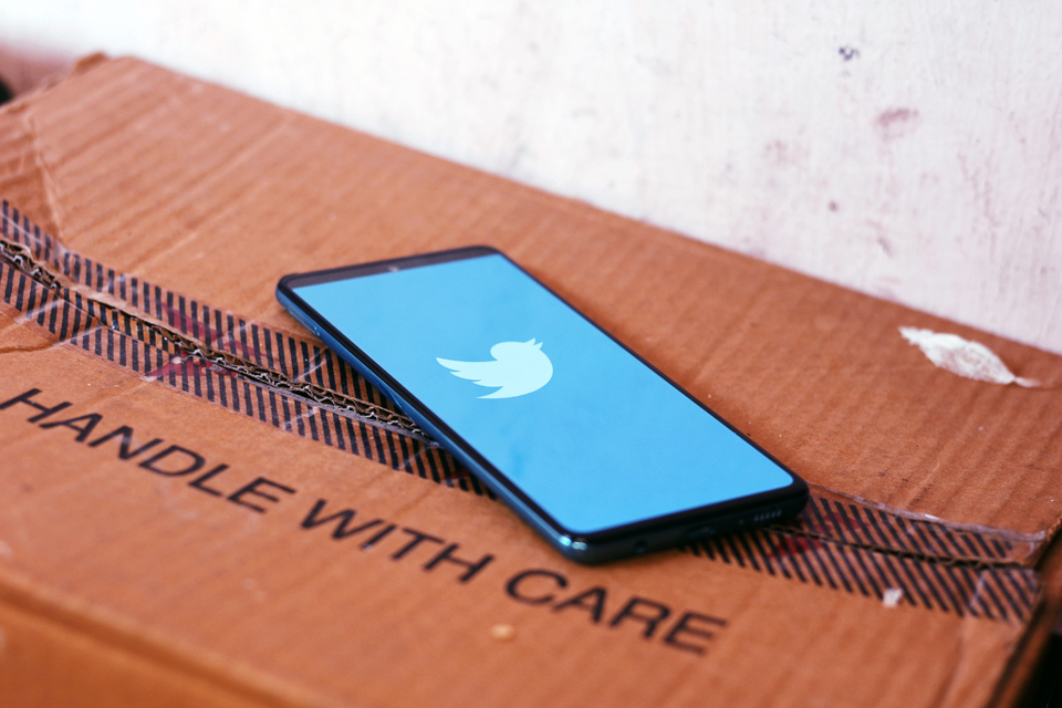 Black tablet showing the blue and white Twitter logo on top of a cardboard box with 'handle with care' stamped on a lid flap. By Ravi Sharma, via Unsplash.