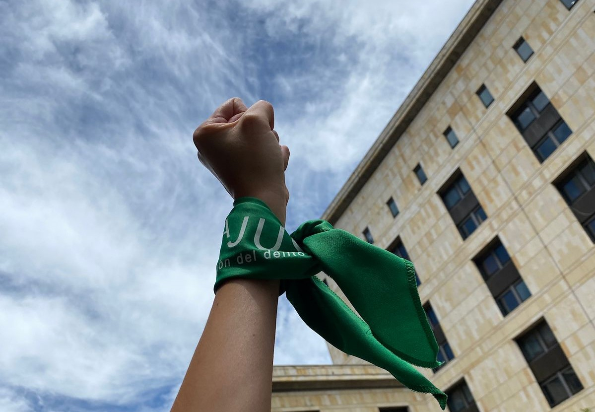 A white fist raised in the air with a green scarf wrapped at the wrist. You can see the blue sky in the background. 