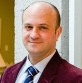 A white man wearing a burgundy blazer and a blue tie smiling at the camera 