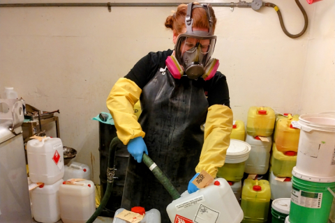 Woman from the HWM team is holding a tube into a chemical waste container. She is wearing a full-face respirator mask, leather apron and long gloves.