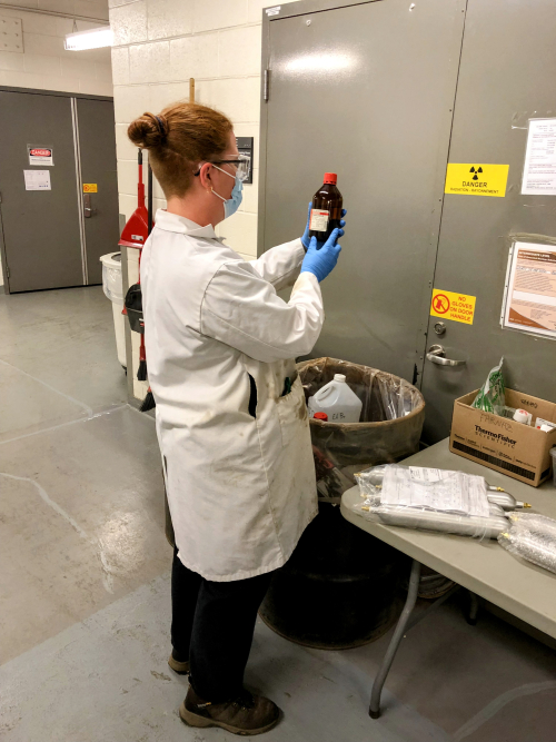 A woman wearing a lab coat, safety goggles, gloves and a mask is looking at a label on a glass bottle that she is holding up. She stands in front of a black steel drum.