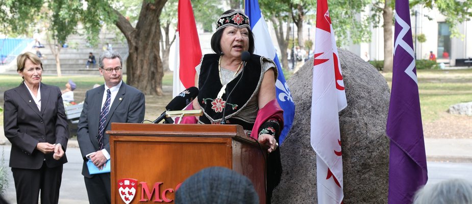 An Indigenous woman in regalia speaks from a podium at an outdoor event at McGill