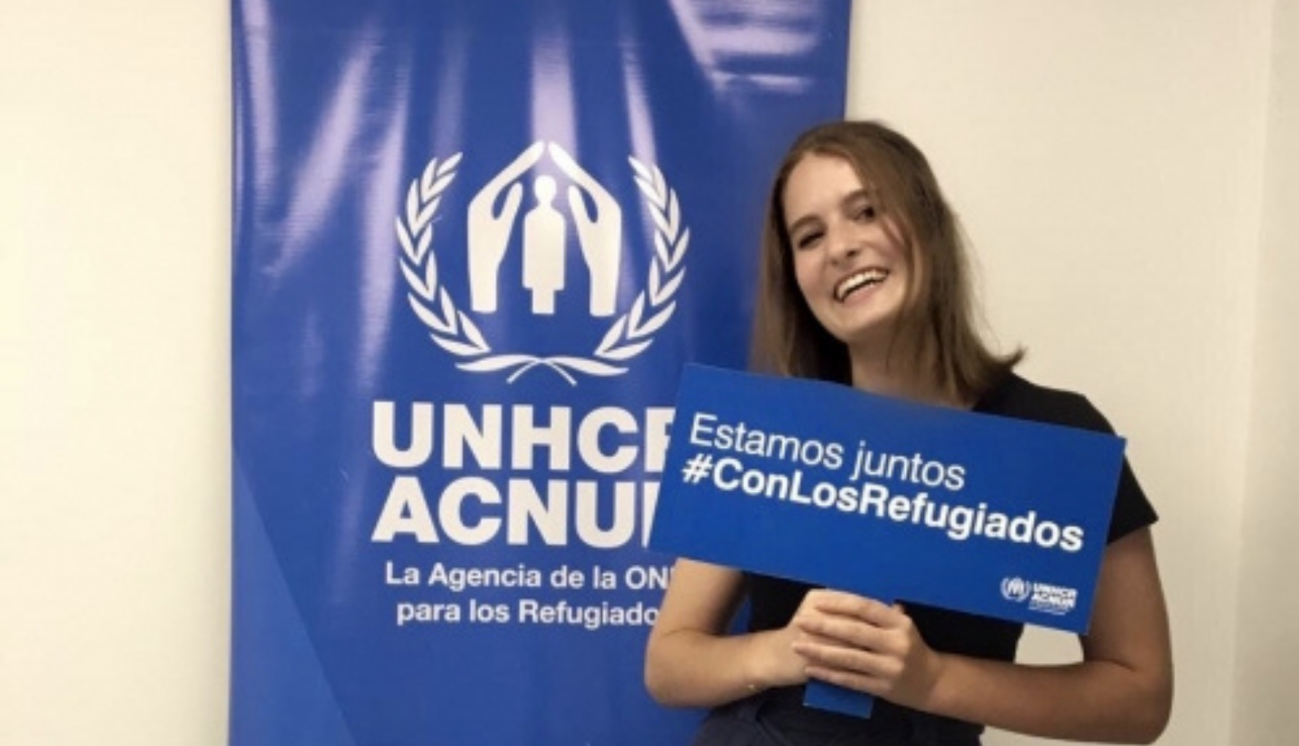 Julia standing in front of the UNHCR logo, holding a sign saying "we are with the refugees" in Spanish