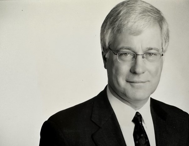 An older white man with short white hair, he is wearing glasses and a black suit and tie