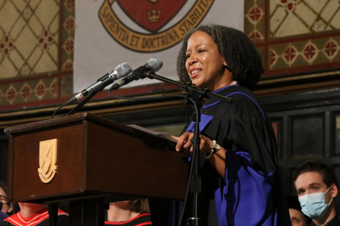 Professor Adelle Blackett (a middle aged black woman with shoulder length hair) standing at a podium wearing a black and blue gown. 
