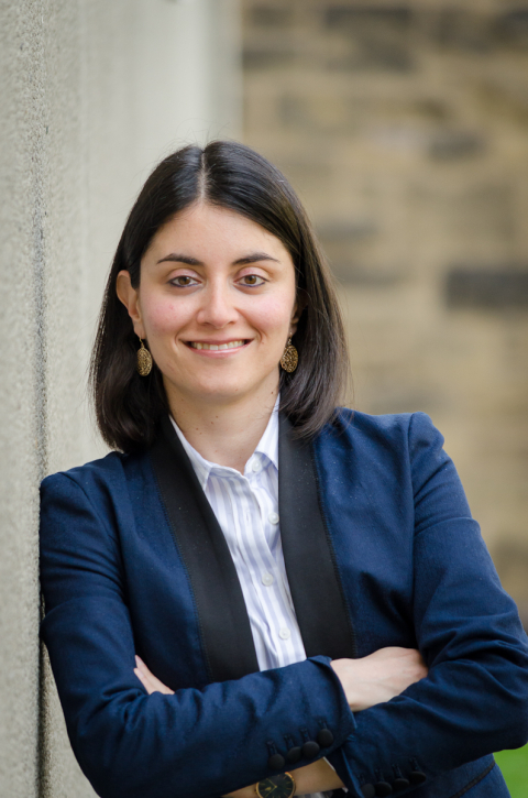 A white woman with shoulder length black hair leaning against the wall and smiling. She is wearing a blue shirt and a navy blazer. 
