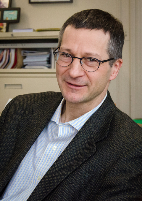 A white man with short black hair and glasses wearing a blue shirt with a black blazer 