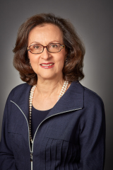 An elder white woman with brown eyes and brown glasses. She has wavy brown hair and is wearing a denim cardigan with pearls.