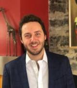 A white man with short brown hair and a suit smiling standing in a dining room.