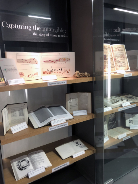 Installation view of the exhibition featuring books, scores, and papers on wood shelves behind a glass case.