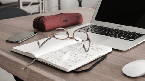 Desk with an open notebook, glasses, a pencil case, a mobile phone, and an open laptop.