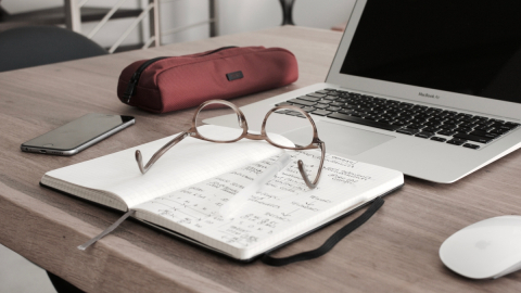 Laptop on desk with glasses, a notebook and a pencil case.