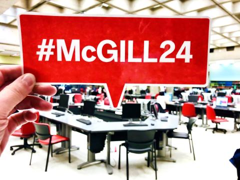 Close-up of hand holding a red #McGill24 cut-out in front of a Library study space.