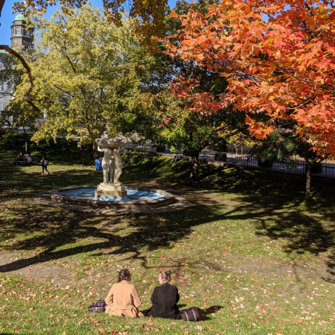 Two people sitting in front of the Three Bares statue surrounded by trees displaying fall colours.