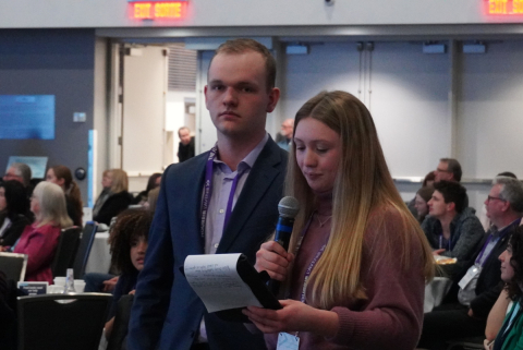 Students Beatrice Neveu and Connor Velthuis stand in front of conference goers with a microphone.